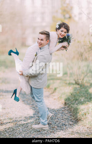Beautiful happy smiling wedding couple with groom lifting up his charming bride in stylish blue heels at the spring park Stock Photo