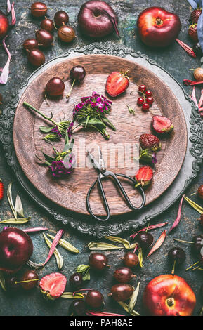 Wooden plate with seasonal fruits, berries, flowers and scissors on rustic kitchen table background, top view. Country style Stock Photo