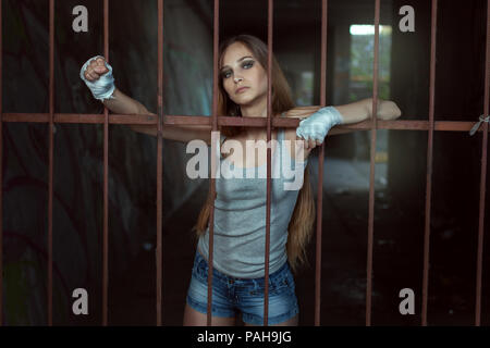 Young female fighter stands behind an iron fence and put her hands on him. Stock Photo