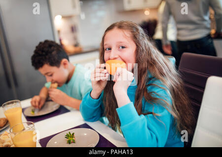 Little girl is looking at the camera while eating a slice of orange at home with her brother. Stock Photo