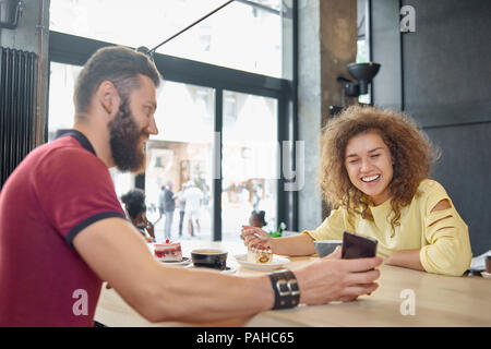Laughing couple eating cake, drinking coffee, looking at smartphone. Male model wearing burgundy polo shirt, brown beard, curly girl having yellow blouse, beatiful smile. Stylish loft design. Stock Photo