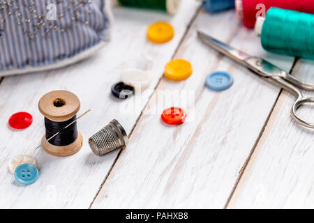 Bright sewing spools with vibrant colorful threads and assorted buttons scattered across rough light wooden plank table with vintage scissors, thimble Stock Photo