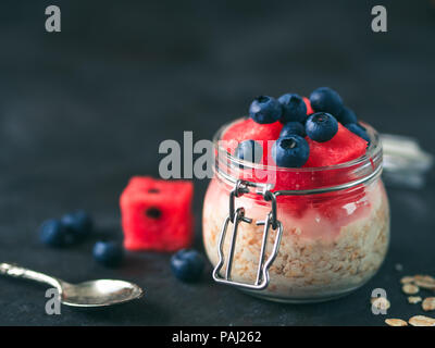 Healthy breakfast idea and recipe: overnight oats with fresh fruits and berries in glass jar.Overnight oatmeal porridge with watermelon,blueberry.Overnight oats on black background.Copy space for text Stock Photo