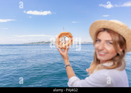 Beautiful woman traveler holds traditional Turkish street food simit(bagel) in background of Maiden Tower,a popular destination in Istanbul,Turkey Stock Photo