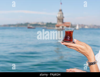 Beautiful woman traveler holds traditional Turkish tea glass in background of Maiden Tower,a popular destination in Istanbul,Turkey Stock Photo