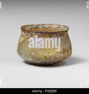 Glass cup. Culture: Roman. Dimensions: Height: 2 3/8in. (6.1cm)  Diam.: 3 3/8 x 2 7/8 in. (8.6 x 7.3 cm). Date: 2nd-3rd century A.D..  Uncertain, probably colorless with pale blue green tinge.  Knocked-off, uneven rim; slightly bulging collar below rim; sides expanding downward, then angled in to join bottom with uneven pushed-in center.  A single wheel-abraded horizontal line around neck below collar and a band of faint lines on body above angle.  Intact; many pinprick bubbles; dulling, deep pitting, and brilliant iridescent weathering on exterior; thick creamy weathering on interior.  Stands Stock Photo