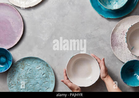 Female hands hold a white ceramic bowl on a gray marble table. Clay handcraft bowls, plates of different sizes are empty. Stock Photo