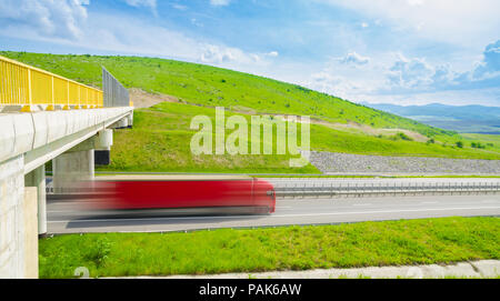 Speeding fast red truck on a highway over the green hills on a sunny summer day with a bright blue cloudy sky and a bridge with a yellow railing sugge Stock Photo