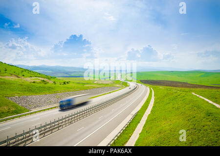 Speeding shipping transport truck on a highway waving over the hills with a fast quick motion look on a sunny day with green grass and a blue cloudy s Stock Photo