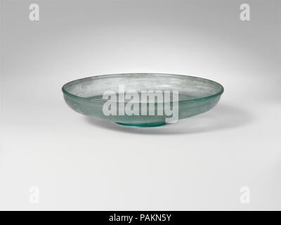 Glass dish. Culture: Roman. Dimensions: H. 1 1/4 in. (3.2 cm)  diameter  5 5/8 in. (14.3 cm). Date: 4th century A.D..  Translucent blue green.  Vertical, slightly thickened, rounded rim; short, convex side to body,  turned in horizontally, and then sloping inwards to thick, flat bottom with upward kick inside and central pontil scar, surrounded by tubular base ring.   Intact; many pinprick bubbles and blowing striations; dulling and faint iridescence. Museum: Metropolitan Museum of Art, New York, USA. Stock Photo