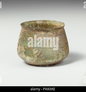 Glass cup. Culture: Roman. Dimensions: Overall: 2 3/4in. (6.9cm)  Diam.: 3 11/16 x 3 1/8 in. (9.4 x 7.9 cm). Date: 2nd-3rd century A.D..  Colorless with blue green tinge.  Knocked-off, uneven rim; slightly bulging collar below rim; sides expanding downward, then angled in to join bottom with pushed-in center.  Band of faint wheel-abraded horizontal lines on body above angle.  Intact; many pinprick bubbles; dulling, deep pitting, and brilliant iridescent weathering on exterior; only faint weathering on interior. Museum: Metropolitan Museum of Art, New York, USA. Stock Photo