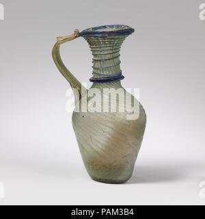 Glass jug. Culture: Roman, Syrian. Dimensions: H.: 4 11/16 in. (11.9 cm). Date: 4th-6th century A.D..  Translucent blue green; handle in same color; trails in translucent cobalt blue.  Plain rim, with downward flange on one side; flaring mouth; cylindrical neck, expanding slightly downwards; slanting, rounded shoulder; funnel-shaped body;  thick bottom with kick and small, central pontil scar; reeded strap handle attached unevenly to shoulder, drawn up and outwards in a curve, then attached to edge of rim and trailed back on itself.  Narrow ribs extend in a tight spiral down from left to right Stock Photo