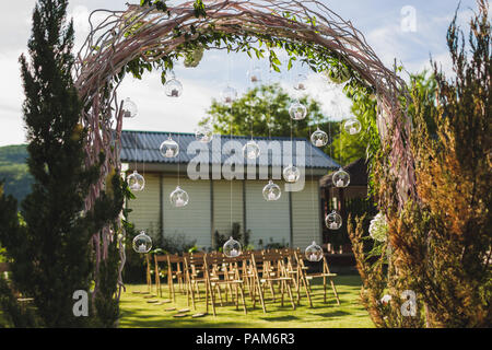 Evening wedding ceremony in garden, arch with white branches, brown wooden chairs and a lot of lights hanging in glass spheres Stock Photo