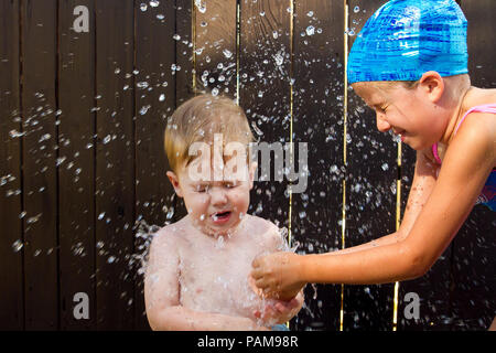 A young girl wearing a blue swim cap, closes her eyes as she bursts a water balloon in the face of her little brother.  Water droplets fly through the