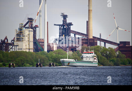 Seagoing cargo ship on a river with green foliage, huge steel mill and several wind power stations in the background