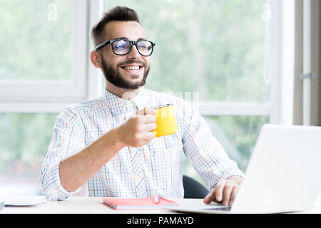 Portrait of trendy bearded businessman enjoying cup of coffee  and smiling happily while working at desk in office using laptop, copy space Stock Photo
