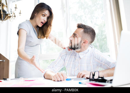 Portrait of agitated businesswoman looking shocked while explaining something to handsome colleague sitting at desk in office Stock Photo