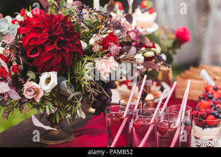 Wedding reception table decorated with red flowers, sweets, drinks and silk. Rustic style Stock Photo