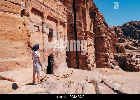 a tourist poses on a rock ledge looking over a tomb entrance in the Lost City of Petra, Jordan Stock Photo