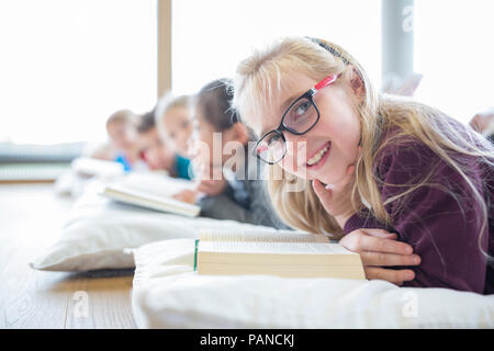 Portrait of smiling schoolgirl lying on the floor with classmates reading book in school break room Stock Photo