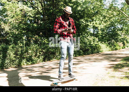 Cool young man skateboarding in park, using smartphone Stock Photo
