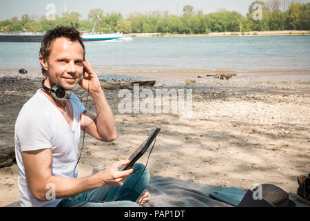 Portrait of smiling man sitting on blanket at a river using tablet Stock Photo
