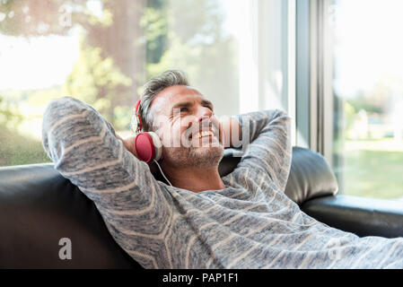 Happy mature man lying on couch at home wearing headphones Stock Photo