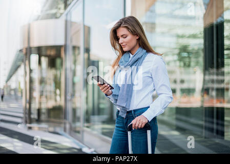 Young businesswoman with suitcase looking at smartphone Stock Photo