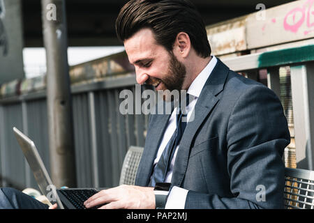 Happy businessman sitting outdoors on a bench using laptop Stock Photo