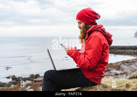 Iceland, North of Iceland, man with laptop sitting near the sea using cell phone Stock Photo
