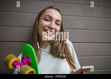 Portrait of smiling teenage girl with cell phone, earphones and skateboard Stock Photo