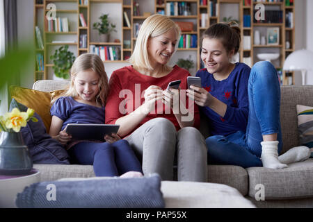 Mother and her daughters sitting on couch, having fun  using digital laptop and playing with smartphones Stock Photo