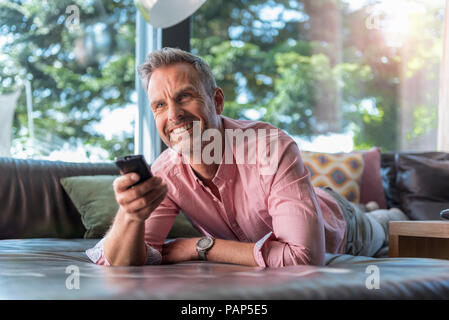 Smiling mature man lying on couch at home using remote control Stock Photo