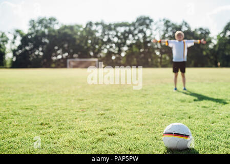 Boy with outstretched arms standing on soccer field between German soccer ball and goal Stock Photo