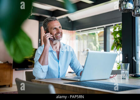 Smiling mature man at home using cell phone and laptop at table Stock Photo
