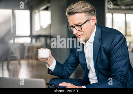 Mature businessman with cup of coffee and laptop using cell phone Stock Photo