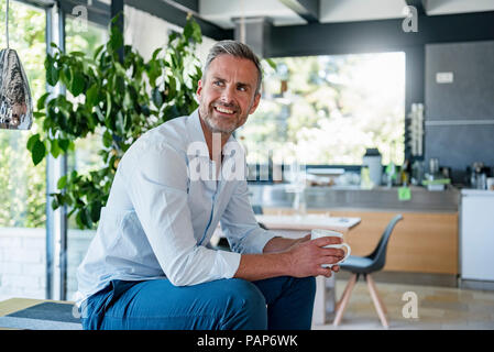 Smiling mature man at home sitting on table with cup of coffee Stock Photo