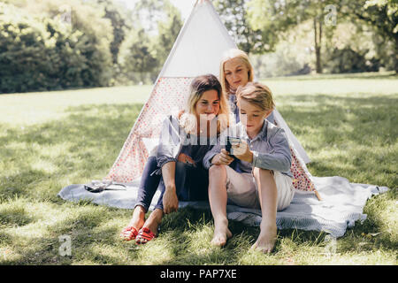 Two young women and a boy looking at cell phone next to teepee in a park Stock Photo