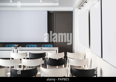 Comfortable conference hall with white walls. There are many light and dark chairs, board, tables with blue soft chairs, lamps. Horizontal. Stock Photo