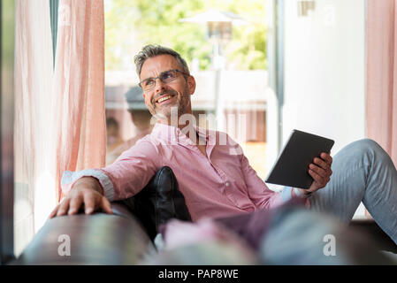 Smiling mature man sitting on couch at home holding a tablet Stock Photo
