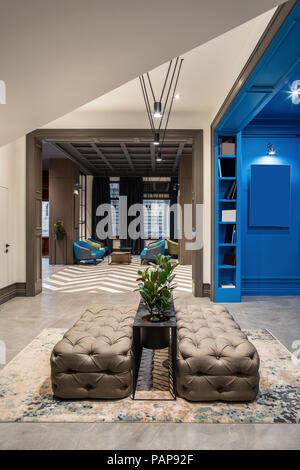 Wonderful hallway in the hotel with blue and light walls and a tiled floor. There are two sofas, shelves with books, black stand with green plants in  Stock Photo