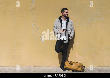 Smiling bearded man with coffee to go, leaning on wall Stock Photo