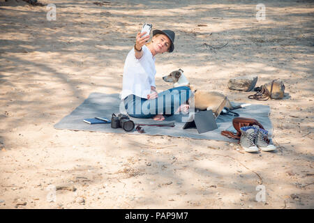 Woman sitting on blanket on beach with dog taking a selfie Stock Photo