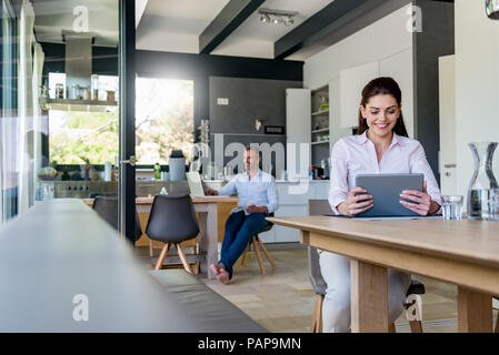 Smiling woman at home using a tablet at table with man in background using laptop Stock Photo