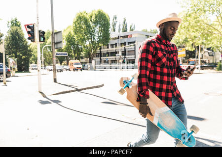 Cool young man with skateboard walking in the city, using smartphone Stock Photo