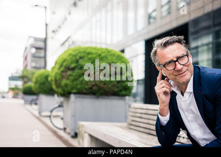 Smiling mature businessman sitting on bench outdoors wearing earbuds Stock Photo