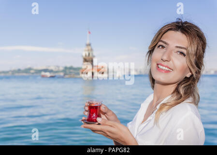 Beautiful woman traveler holds traditional Turkish tea glass in background of Maiden Tower,a popular destination in Istanbul,Turkey Stock Photo