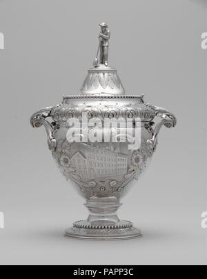 Sugar bowl. Culture: American. Dimensions: 9 1/16 x 6 1/4 in. (23 x 15.9 cm). Maker: Wood and Hughes (1845-99). Date: 1862-63.  Presented to Christian Dorflinger upon his retirement from the Long Island Flint Glass Works in 1863, this service (2006.90.1-4) is ornamented with meticulously chased scenes of the glassworks itself. The sugar bowl is ornamented with an image of the exterior of the factory building. The finials, cast in the form of glassblowers, also reference Dorflinger's trade. Dorflinger was one of the most successful nineteenth-century American glassmakers, operating several fact Stock Photo