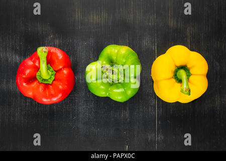Top view of three fresh bright bell peppers on black rustic background. Shot from above of green, yellow and red paprika vegetables on dark wood table Stock Photo