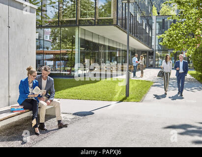 Smiling colleagues with book sitting on bench outside office building Stock Photo